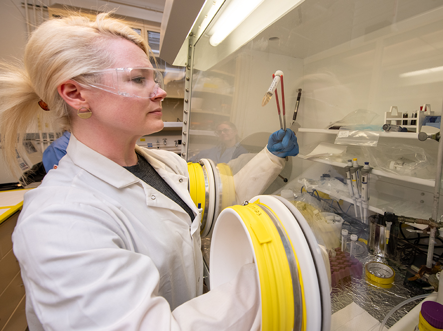 Researcher in white coat and safety glasses studying sample behind safety glass