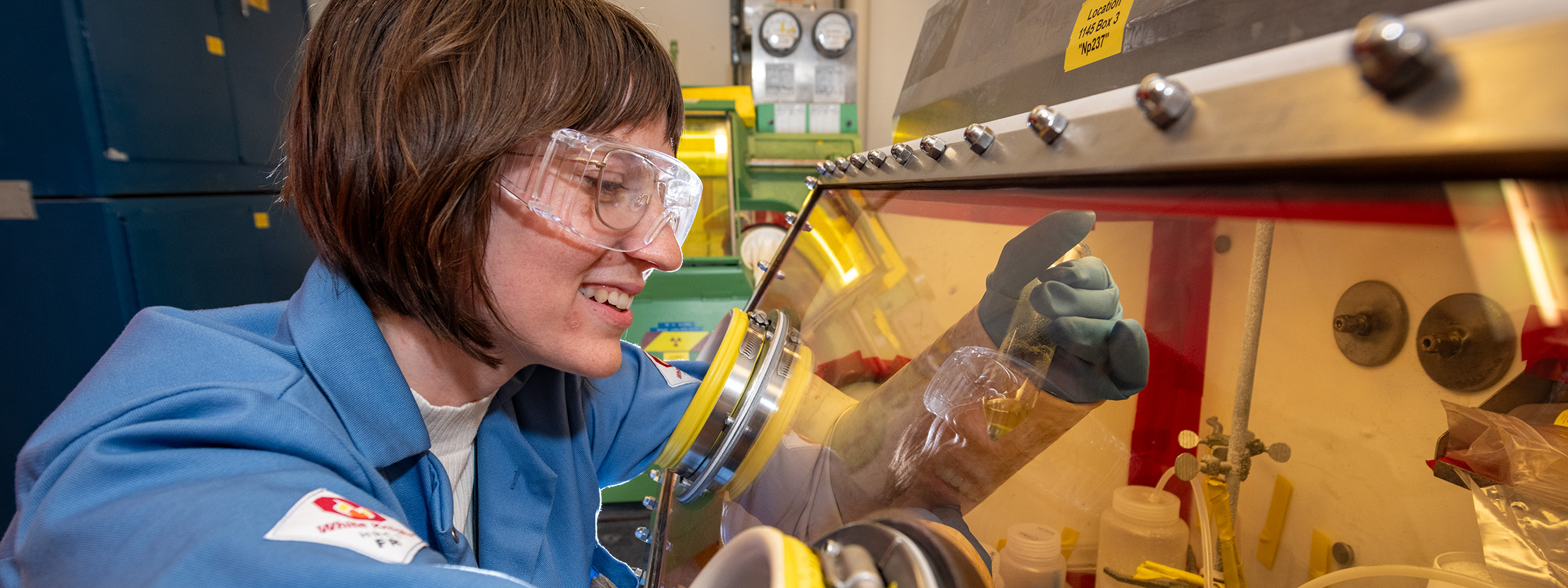 Researcher with safety goggles on leans over studying sample behind safety glass