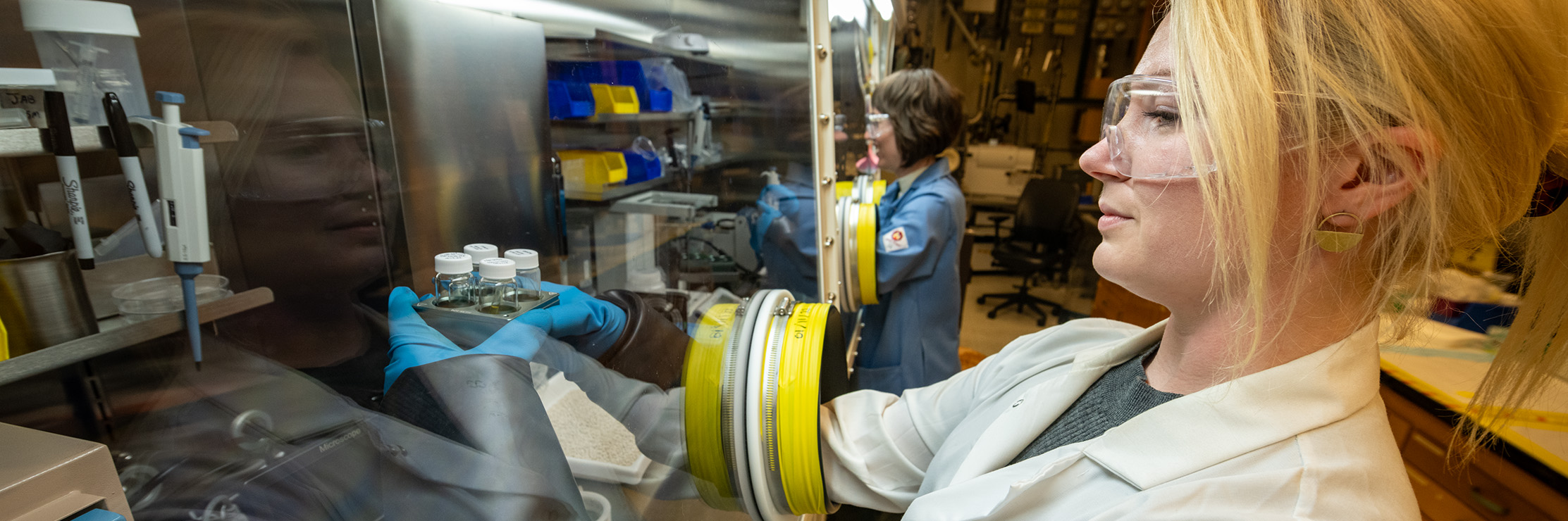 research in foreground looking at scientific samples behind safety glass.